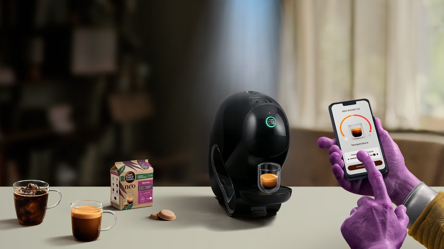 A mão de um extraterrestre a utilizar a aplicação NESCAFÉ Dolce Gusto no telemóvel para personalizar o seu café. Máquina NEO, embalagens de café e bebidas no fundo.