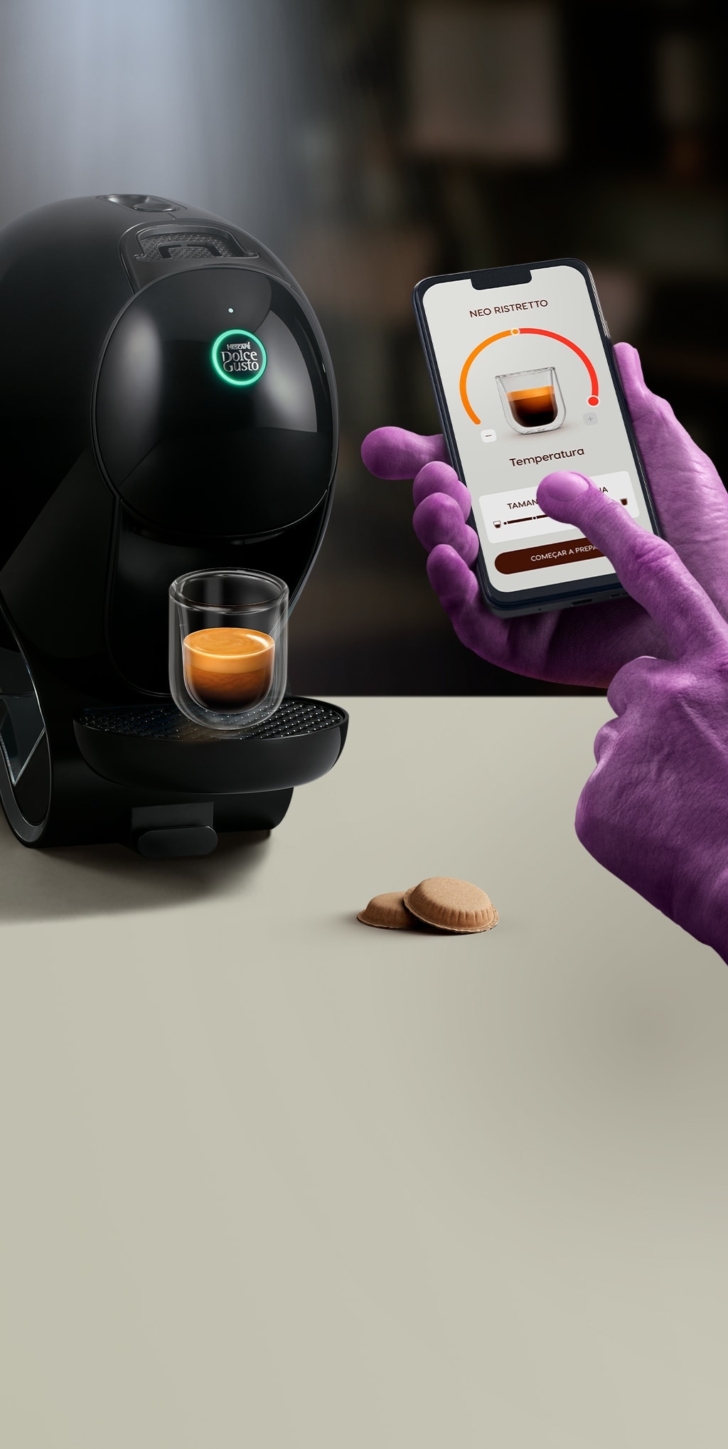 A mão de um extraterrestre a utilizar a aplicação NESCAFÉ Dolce Gusto no telemóvel para personalizar o seu café. Máquina NEO, embalagens de café e bebidas no fundo.